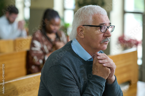 Mature male parishioner with grey hair praying with his hands put together while sitting on bench against other people