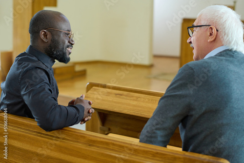 Young African American pastor consulting senior male parishioner after church service while both sitting on bench in front of one another photo