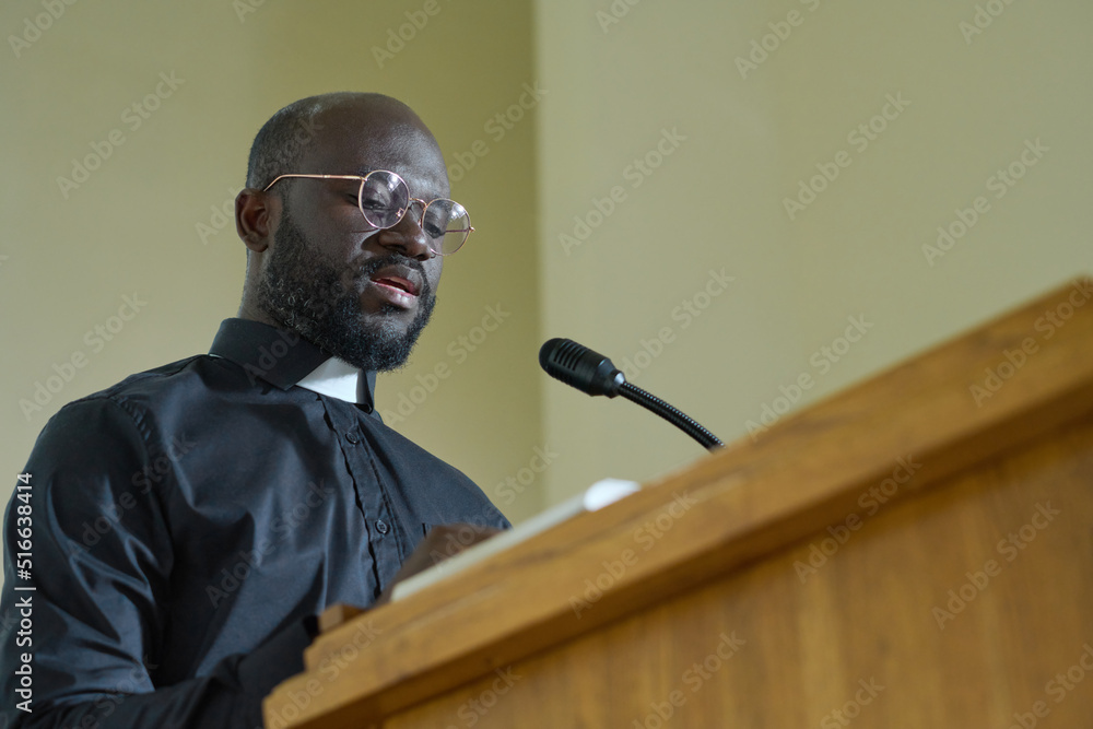 Young priest in black shirt with clerical collar reading verses from Bible and explaining them while pronouncing speech by pulpit - obrazy, fototapety, plakaty 