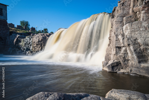 Waterfall with quartzite red rocks at Big Sioux River Park in Sioux Falls  South Dakota. Long exposure photo of the silky flowing water with the view of the Queen Bee Turbine House on the hilltop.