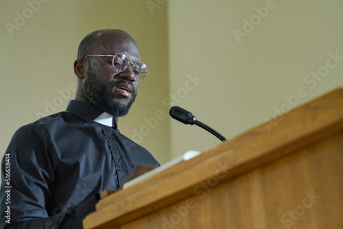 Young priest in black shirt with clerical collar reading verses from Bible and explaining them while pronouncing speech by pulpit photo