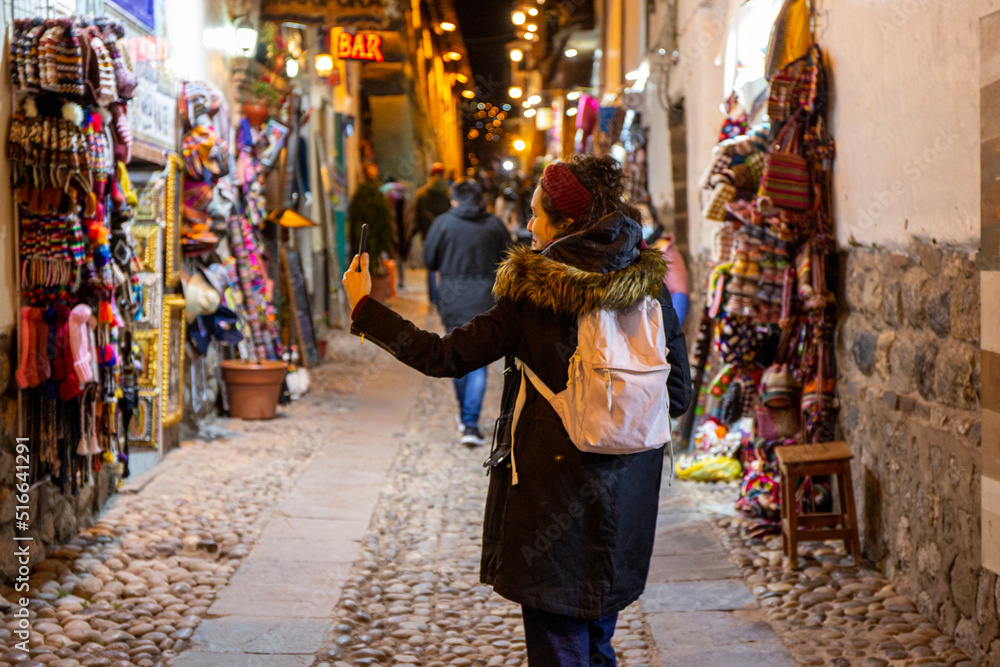 Tourist woman walking through Cusco street at night between shops with cell phone in hand