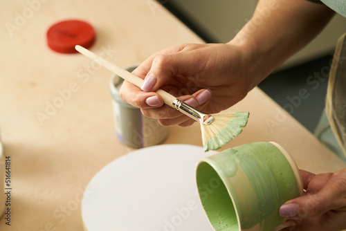 Close-up of girl painting clay mug with glaze. Woman coloring pottery in workshop with a paintbrush. Painter in green apron glazing clay pot