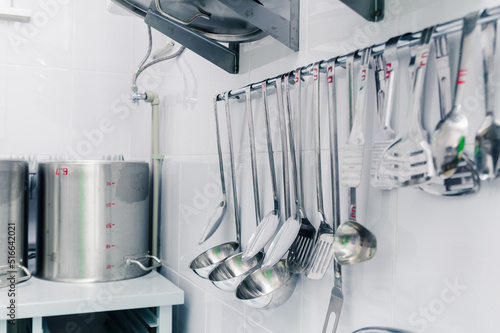 A set of basic kitchen utensils hangs on the wall with an aluminum shelf for many lids photo