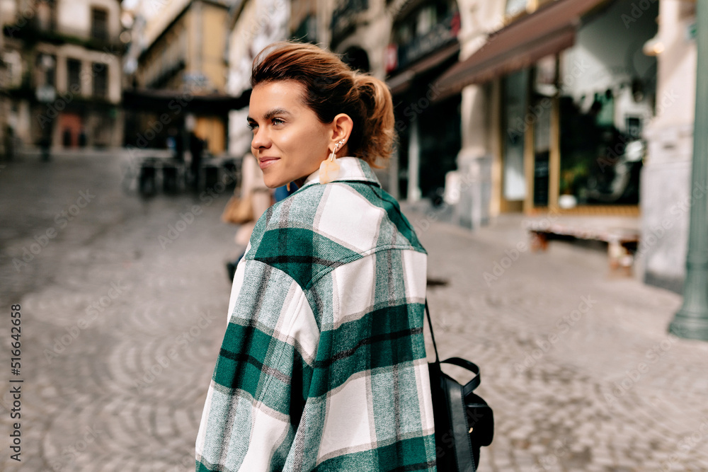 Fototapeta premium Happy smiling lady with collected hair wearing checkered shirt with backpack walking around city in sunny spring day. Photo of fashionable girl with beautiful brown hair