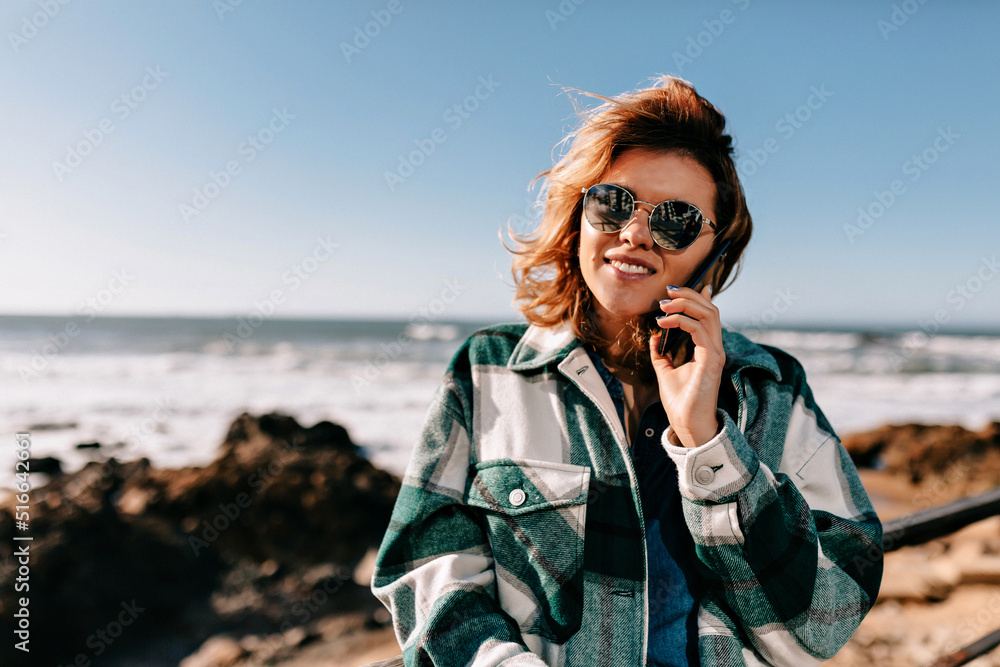 European pretty girl with happy smile talking on the phone in sunny warm day on ocean background. Girl walking around the ocean shore. Stylish woman with smartphone on the ocean European pretty girl with happy smile talking on the phone in sunny warm day on ocean background. Girl walking around the ocean shore. Stylish woman with smartphone on the ocean