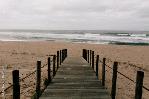 Wooden bridge leads to the ocean waves and sandy clean beach in spring day. Blue sky with clouds and fog. Portugal 