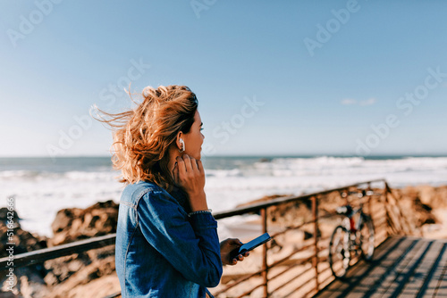 Outdoor portrait from back of european girl with loose hair wearing denim shirt listening music and walking along the ocean