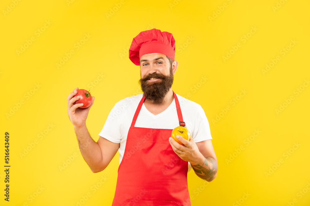 Happy guy in cooking apron and toque holding red and yellow tomatoes yellow background, cook Happy guy in cooking apron and toque holding red and yellow tomatoes yellow background, cook