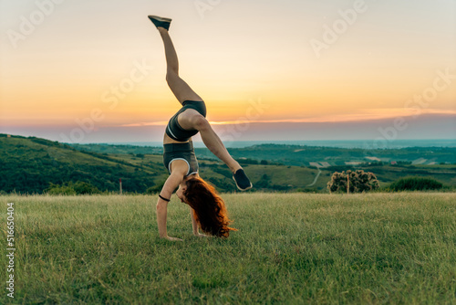 Sporty young woman performing handstand on grass field in sunset.