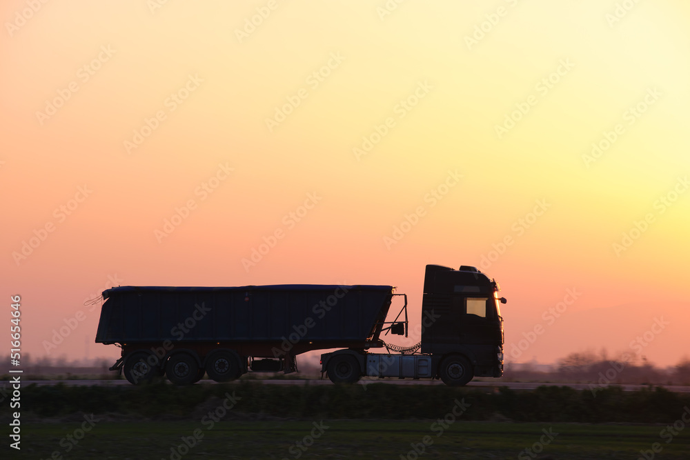 Semi-truck with tipping cargo trailer transporting sand from quarry driving on highway hauling goods in evening. Delivery transportation and logistics concept Semi-truck with tipping cargo trailer transporting sand from quarry driving on highway hauling goods in evening. Delivery transportation and logistics concept