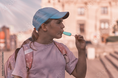 A child of 10-12 years old eats popsicles on a city street. Girl in a cap with a backpack eats ice cream on a hot summer day.