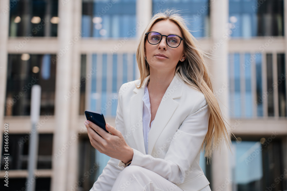 Calm blonde businesswoman in glasses wearing white suite sitting in front of business center looking confidently aside. Successful European lawyer going to call her client. Business and finance.