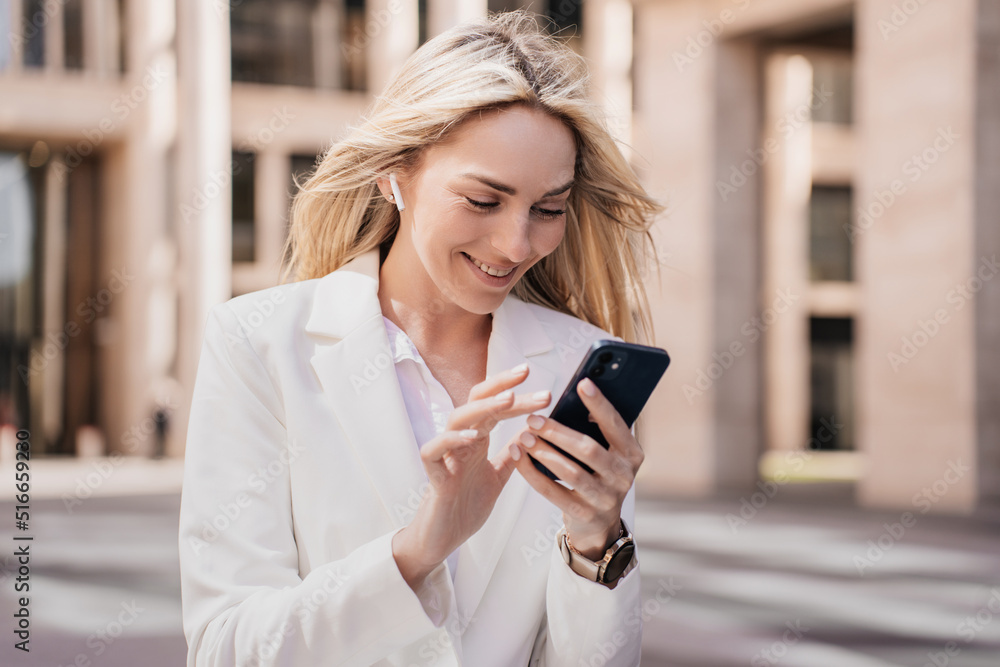 Excited young blonde businesswoman woman making video call by phone, earphones, with happy facial expression smiling sitting outdoors against building. Pretty lawyer typing message. Happy cute student