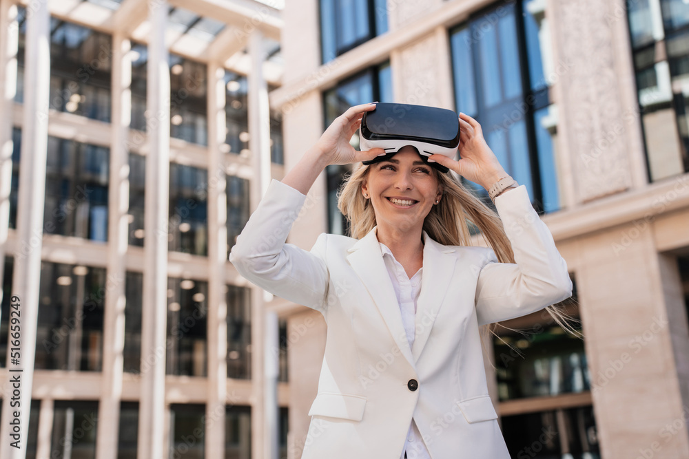 Cheerful Italian woman in white suit using virtual reality glasses in the city. Young entrepreneur swedish woman in putting on vr glasses with happy smile against buildings. Technologies and leisure.