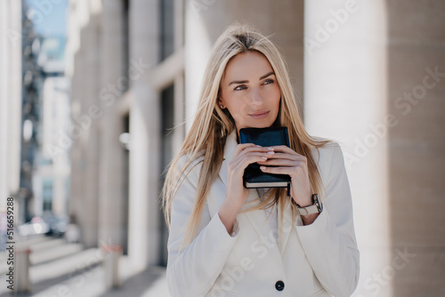Cheerful swedish blonde young woman in white suit looking seriously  holding diary. Beautiful  businesswoman standing outdoor, planning her day. Business and finance. Overloaded student woman in city.