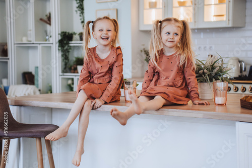Two Scandinavian cute little sisters with ponytails sitting on kitchen table hugging knees looking at camera. Little swedish girls in pyjamas  on breakfast time at home. Family domestic activities. photo