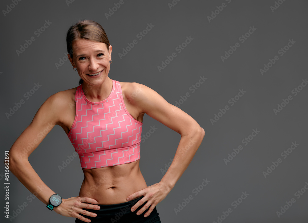 Beautiful woman posing in studio. Portrait of happy young brunette in pink top for yoga or fitness, she stands on gray isolated background and smiles, shows off her athletic figure. Beautiful woman posing in studio. Portrait of happy young brunette in pink top for yoga or fitness, she stands on gray isolated background and smiles, shows off her athletic figure.
