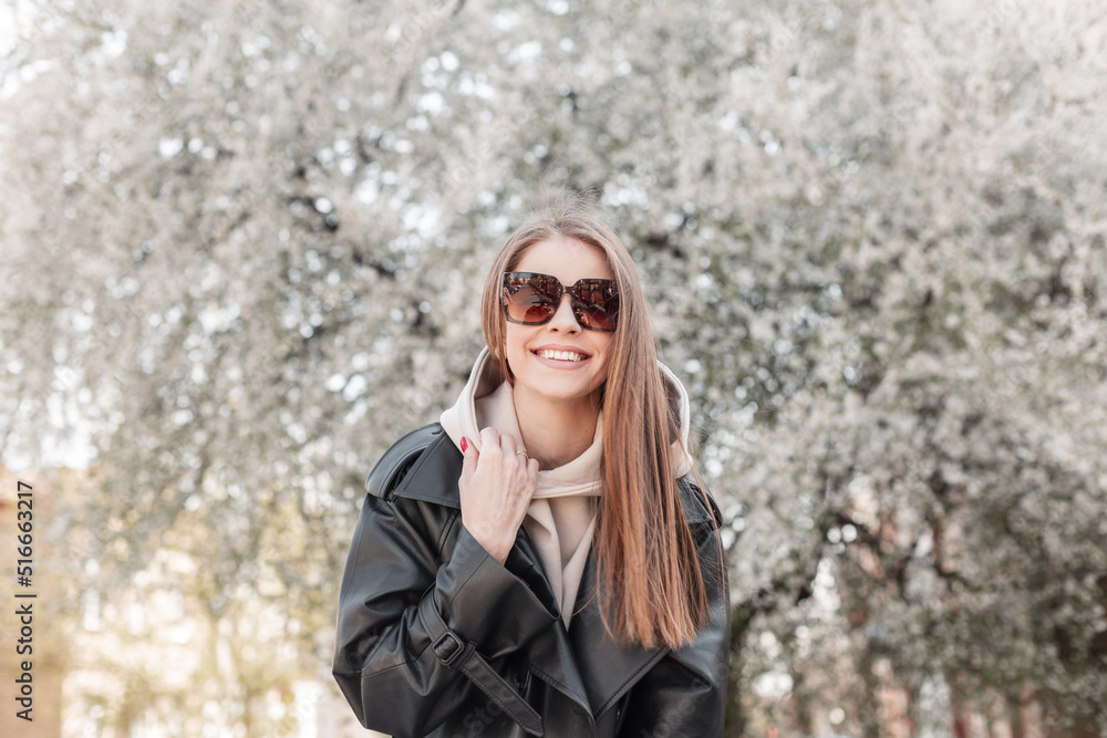 Happy beautiful cheerful fashionable hipster girl with a smile in fashion clothes with a trendy leather jacket, hoodie and sunglasses have fun in a blooming park Happy beautiful cheerful fashionable hipster girl with a smile in fashion clothes with a trendy leather jacket, hoodie and sunglasses have fun in a blooming park