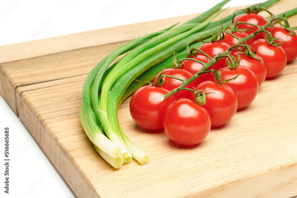 Bunches of fresh red cherry tomatoes and young green onion on cutting board isolated on white background. Ripe tomatoes on green stems. Bunch of fresh onion. Fresh organic vitamin vegetables.