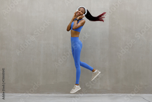 A young woman in headphones with a toned body jumps and runs on a gray background.
