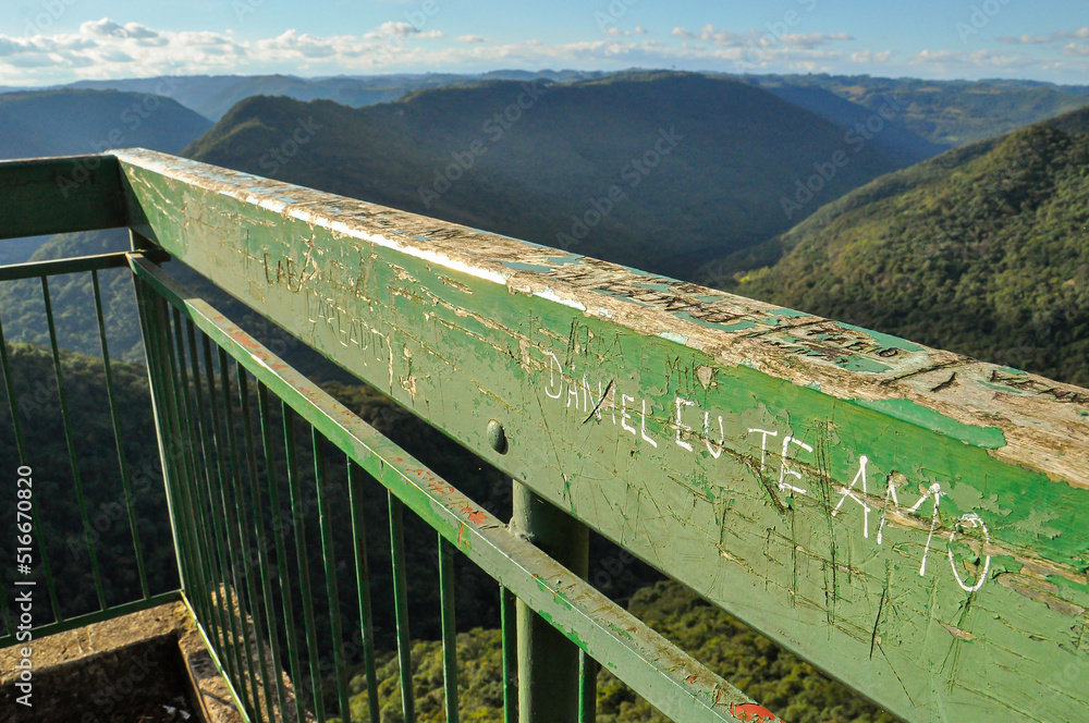 Fototapeta premium Carved markings on the railing of Gelain Lookout, with a scenic view of forested hills and a deep valley in Flores da Cunha, Brazil
