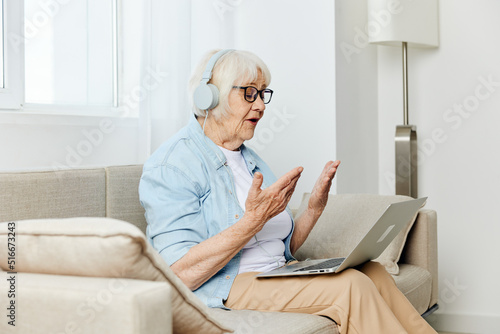 an old woman, carried away by a conversation, is sitting on a cozy sofa and actively communicating via video with headphones on her head while looking at a laptop monitor © Tatiana