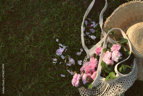 Straw hat and mesh bag with beautiful tea roses on green grass outdoors, flat lay. Space for text