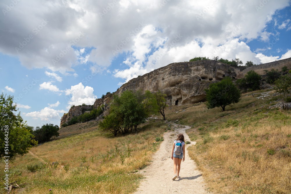 A teenage girl with a backpack climbs a country road towards the stone rocks on a sunny summer day. Travel, tourism concept.