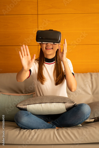 Attractive young woman sits on a sofa chair wearing VR technology equipment glasses and watches a 3D simulated movie making hand gestures.