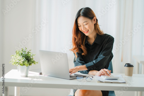 Smiling happy Asian woman relaxing using the technology of a laptop computer while sitting in a chair at a desk. creative girl working at home The concept of working and studying online learning