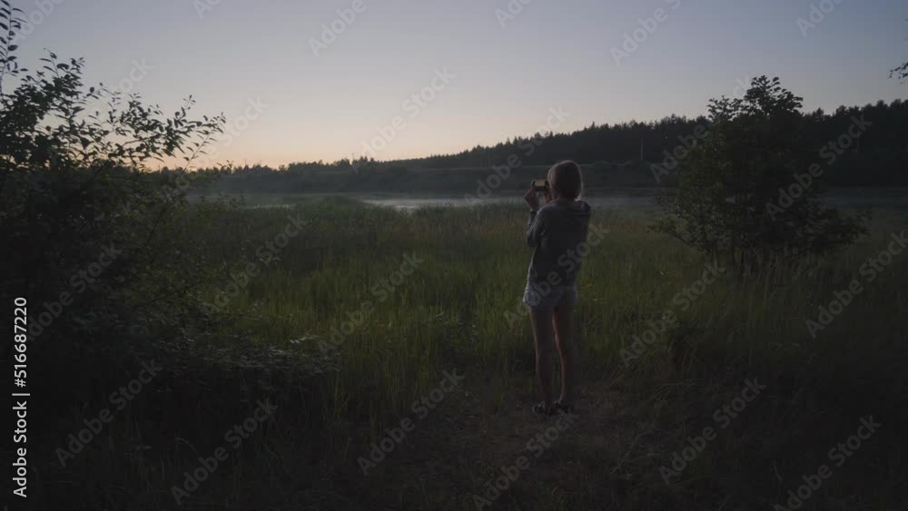 Young Caucasian girl standing taking photos with her smartphone at sunset in the countryside. Young Caucasian girl standing taking photos with her smartphone at sunset in the countryside.