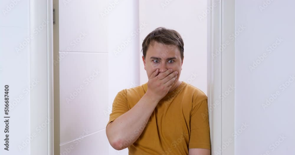 Young man opens door to leave room, but sees something bad in front of him, stops in doorway, leans against door jamb so as not to fall, opens eyes wide and covers mouth with palm in shock Young man opens door to leave room, but sees something bad in front of him, stops in doorway, leans against door jamb so as not to fall, opens eyes wide and covers mouth with palm in shock