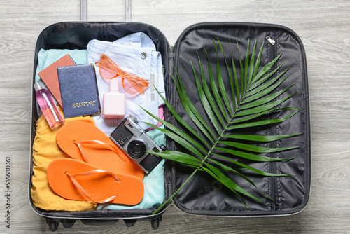Opened suitcase with clothes, accessories for travelling and palm leaf on grey wooden background