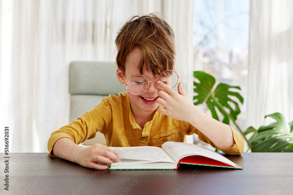 Cute little student in yellow shirt adjusting glasses and smiling while sitting at table and reading book during studies in daytime at home Cute little student in yellow shirt adjusting glasses and smiling while sitting at table and reading book during studies in daytime at home