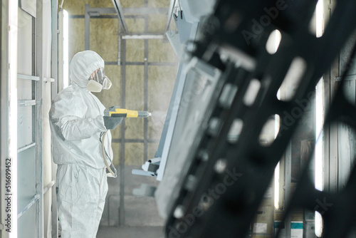 Industrial worker in protective workwear using spray gun to paint metal details at factory photo