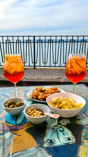 Typical Italian alcoholic aperitif (aperitivo) served in bar in the city center of Ortigia island in the city Syracuse, Sicily, Italy, Europe, EU. Aperol Spritz with ice cubes in summer. Potato chips