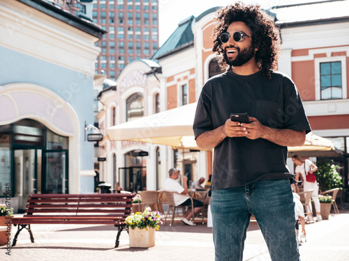 Handsome smiling hipster  model.Sexy unshaven Arabian man dressed in summer clothes. Fashion male with long curly hairstyle using smartphone apps, looking at cellphone screen.Holds phone © halayalex