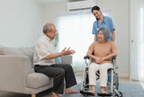 Senior man talking encouraging to his wife patient while sitting on wheelchair and Asian young woman caregiver doctor helps support her during home visit