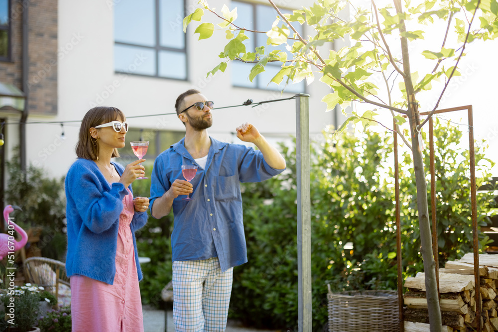 Young stylish couple hang out together and have a drink while standing at their beautiful backyard of country house. Young man and woman spend summer time happily Young stylish couple hang out together and have a drink while standing at their beautiful backyard of country house. Young man and woman spend summer time happily