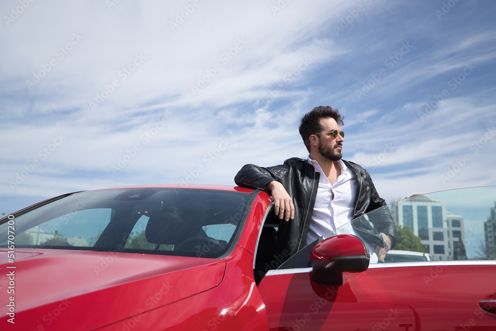 Handsome young man with beard, sunglasses, leather jacket and white shirt, leaning on the roof of his red sports car. Concept beauty, fashion, trend, luxury, motor, sports. Handsome young man with beard, sunglasses, leather jacket and white shirt, leaning on the roof of his red sports car. Concept beauty, fashion, trend, luxury, motor, sports.