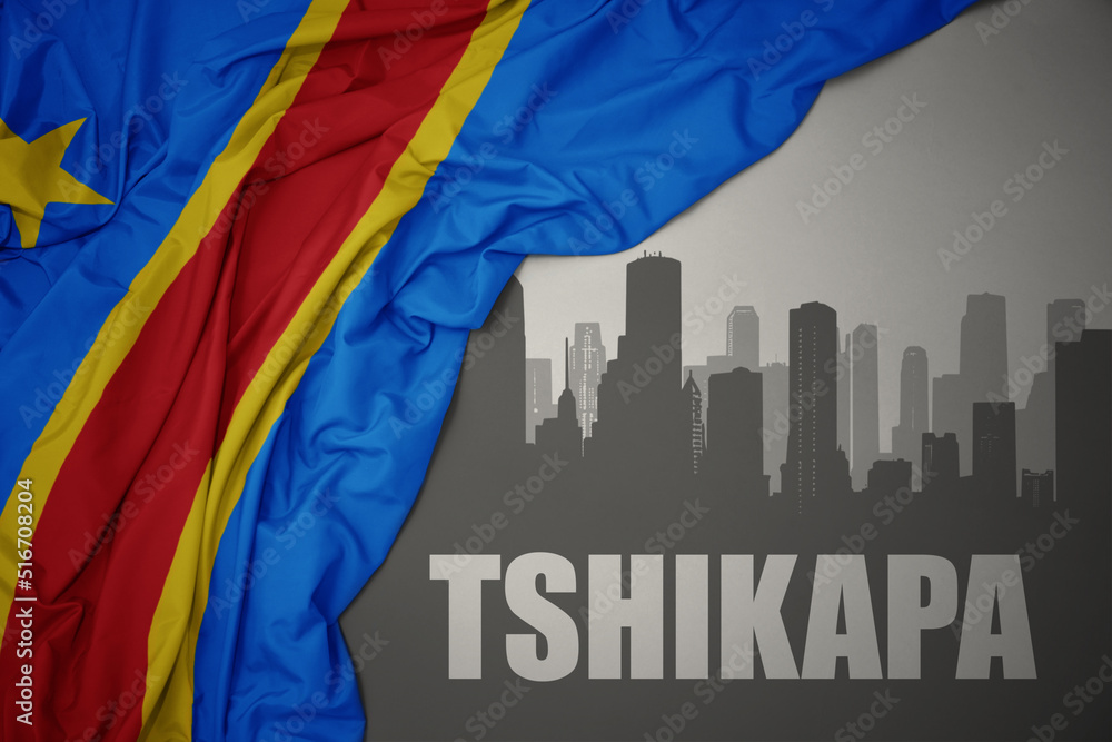 abstract silhouette of the city with text Tshikapa near waving colorful national flag of democratic republic of the congo on a gray background.