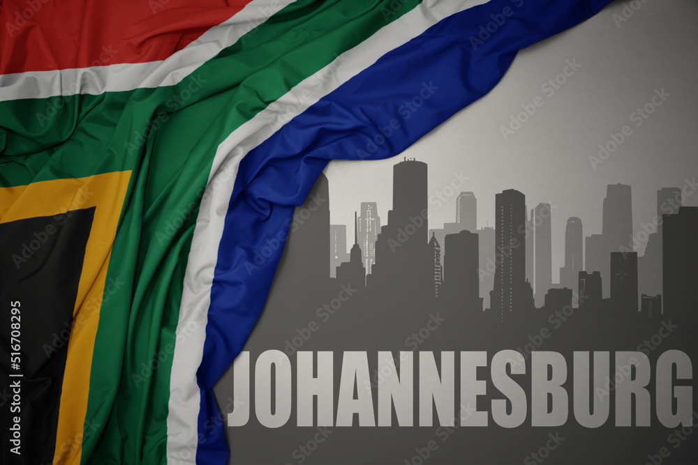 Fototapeta premium abstract silhouette of the city with text Johannesburg near waving colorful national flag of south africa on a gray background.
