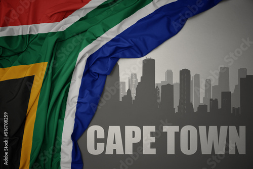 abstract silhouette of the city with text Cape Town near waving colorful national flag of south africa on a gray background.