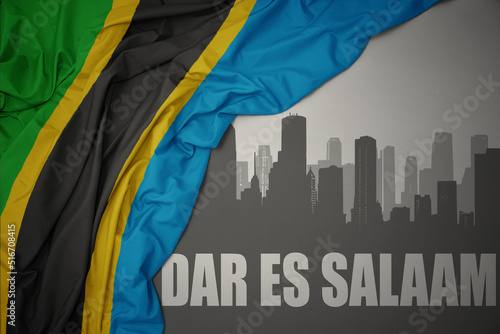 abstract silhouette of the city with text Dar es Salaam near waving colorful national flag of tanzania on a gray background. photo