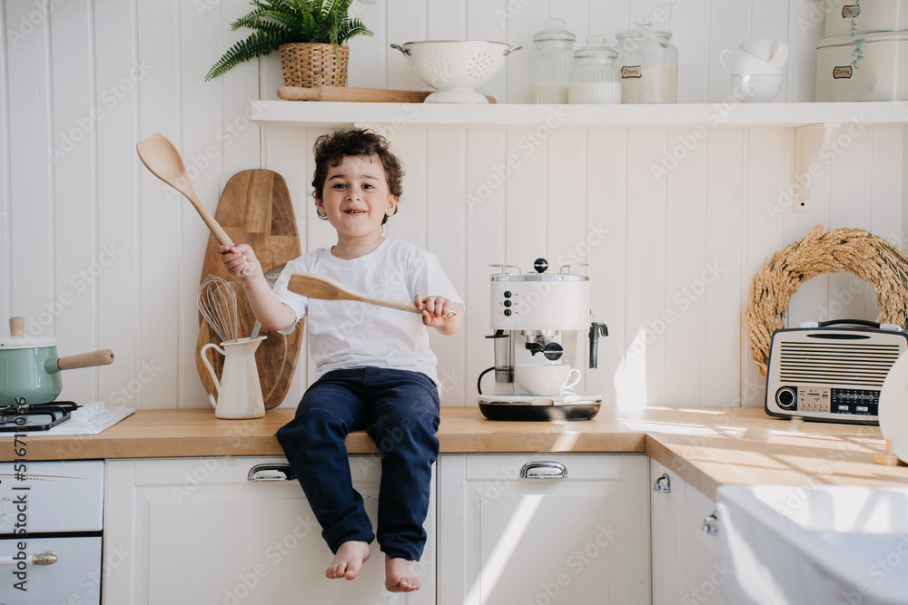 Playful little curly boy in white t-shirt and dark pants laughing sitting on a kitchen table, having fun, misbehaving at home. Spanish boy playing with wooden spoons at kitchen, kitchen stuff around. Playful little curly boy in white t-shirt and dark pants laughing sitting on a kitchen table, having fun, misbehaving at home. Spanish boy playing with wooden spoons at kitchen, kitchen stuff around.