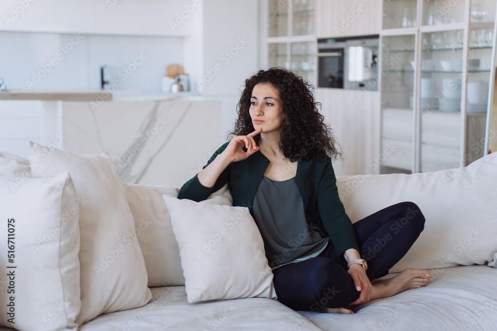 Confident eastern curly female in casual clothes sitting on cozy sofa leaning on cushion looking aside. Thoughtful housewife at living room having rest after housekeeping. Lonely sed woman at home. Confident eastern curly female in casual clothes sitting on cozy sofa leaning on cushion looking aside. Thoughtful housewife at living room having rest after housekeeping. Lonely sed woman at home.