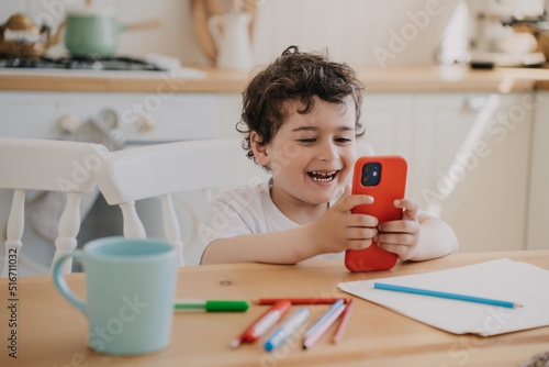 Φωτογραφία Happy cute cheerful curly Spanish  little boy smiling using phone playing games at table at kitchen