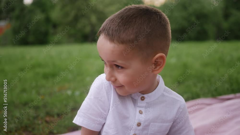 boy child in a white T-shirt sits on a blanket on a green lawn in summer and looks at the camera smiling boy child in a white T-shirt sits on a blanket on a green lawn in summer and looks at the camera smiling