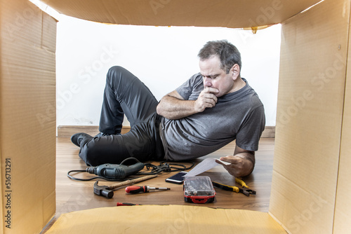 The man is lying on the floor with the tools around and thoughtfully studying the instructions, view of through the open cardboard box photo